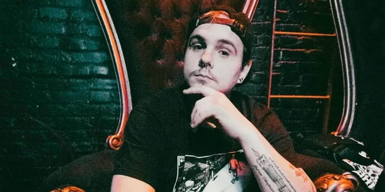 Man with tattoos sitting on an ornate throne-like chair in a dimly lit room.