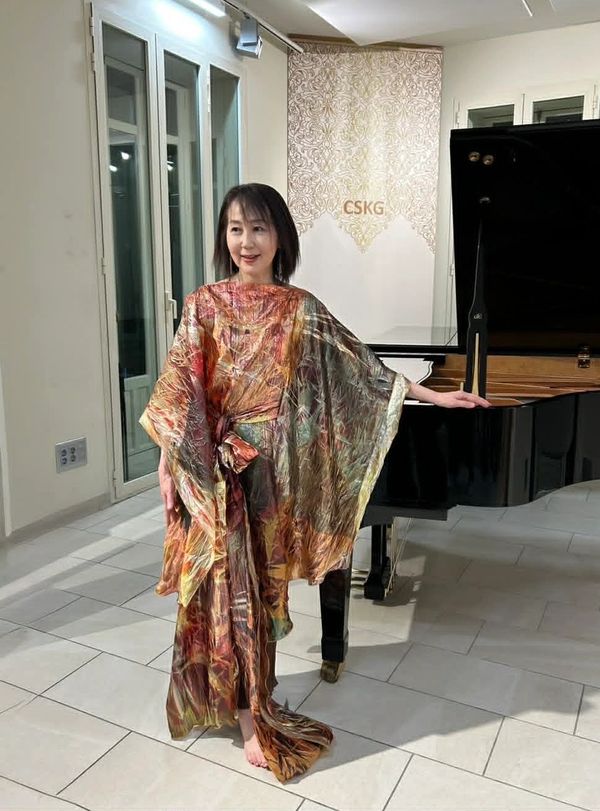 Woman in colorful flowing dress poses by a grand piano indoors.