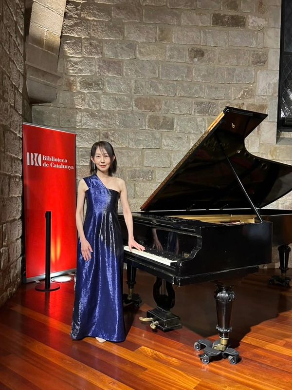 Woman in a blue gown standing beside a grand piano in a stone-walled room.