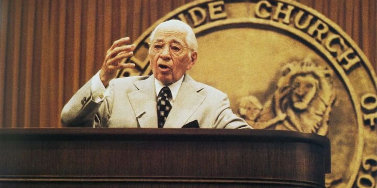 Elderly man delivering a speech at a church podium with a golden emblem behind him.