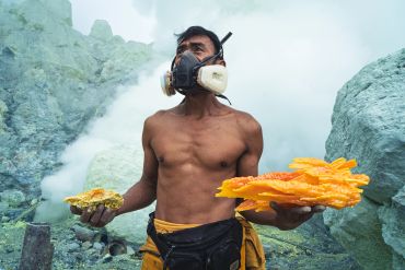 A miner wearing a gas mask holds bright yellow sulfur chunks amid rocky terrain and smoke.