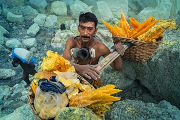 A man carrying baskets of bright yellow sulfur near rocky terrain.