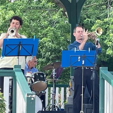 Three musicians perform outdoors under a gazebo with brass and drums.