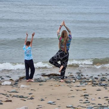 Two people practicing yoga poses on a rocky beach near the ocean.