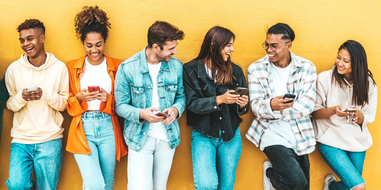 A group of six diverse young adults smiling and using smartphones against a yellow wall.