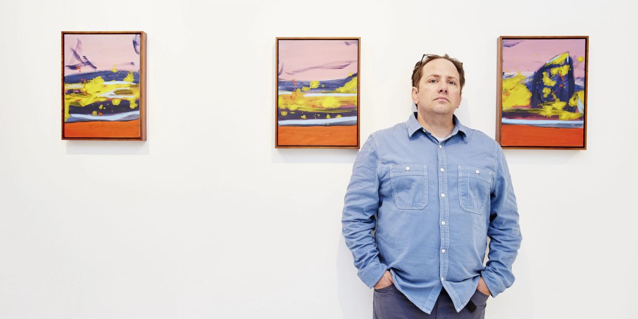 Man in blue shirt stands by colorful abstract paintings on a white wall.
