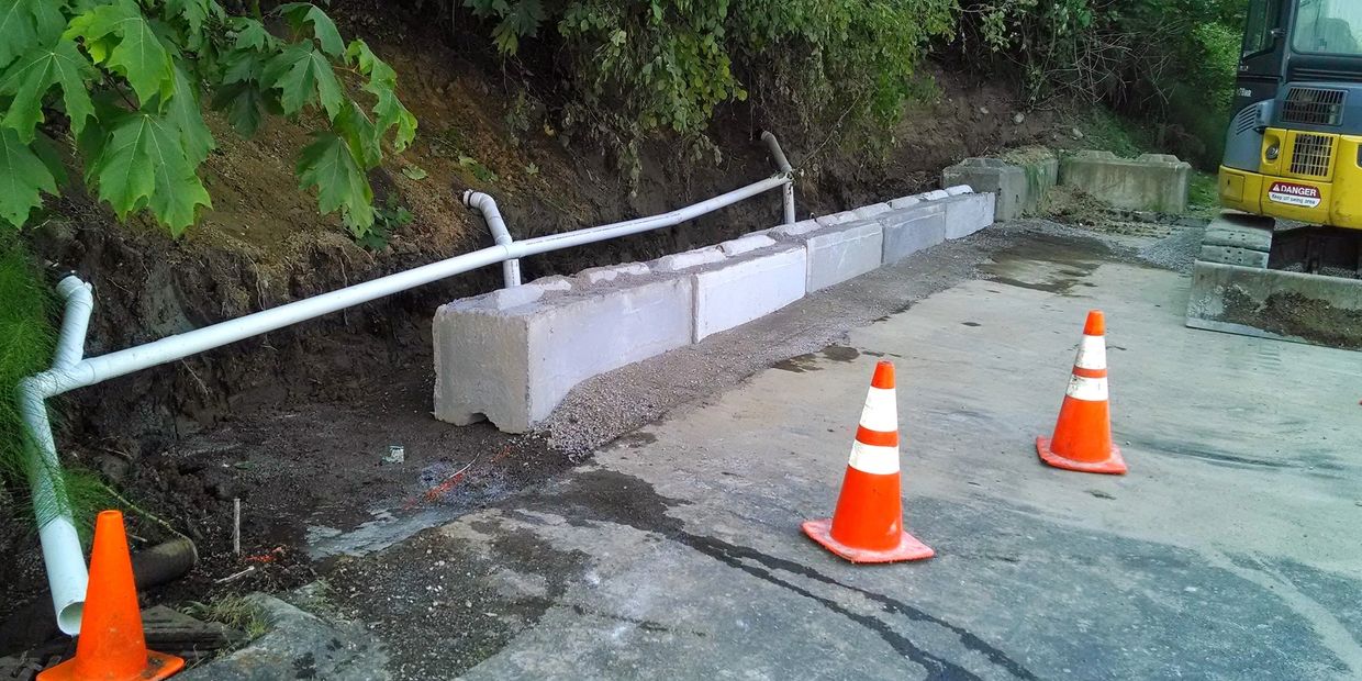 Construction site with concrete blocks, pipes, traffic cones, and an excavator near a wooded area.