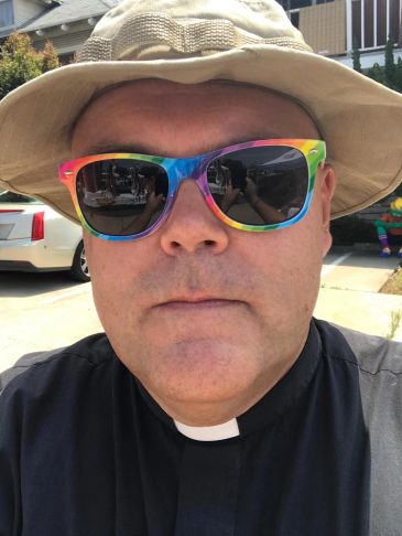 Man wearing rainbow sunglasses and a clerical collar with a sun hat outdoors.