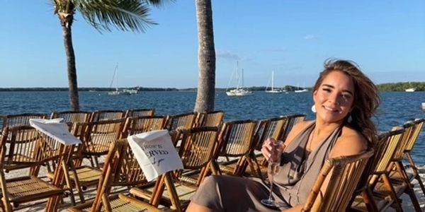 A woman in a gray dress holds a drink on a waterfront with palm trees and boats.
