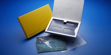 A sleek tech product kit with a yellow box, opened white tray, and photo cards on a blue surface.