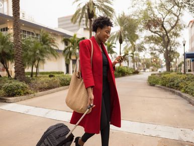 Woman in a red coat walking with luggage and looking at her phone.