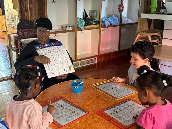 Teacher holding alphabet chart teaching three children at a round table.
