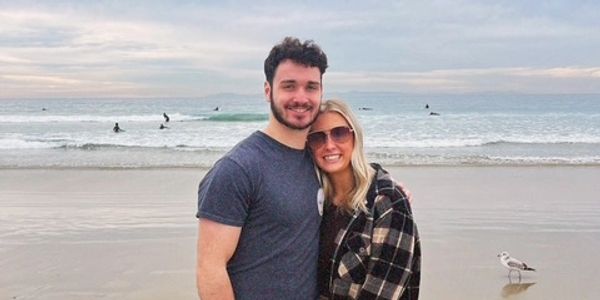 A smiling couple standing close together on a beach with waves and surfers in the background.