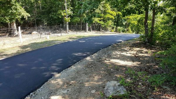 Newly paved asphalt road winding through a wooded area with a wire fence on one side.