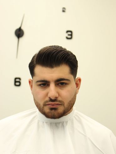 A man with a styled haircut and beard in front of a wall clock.