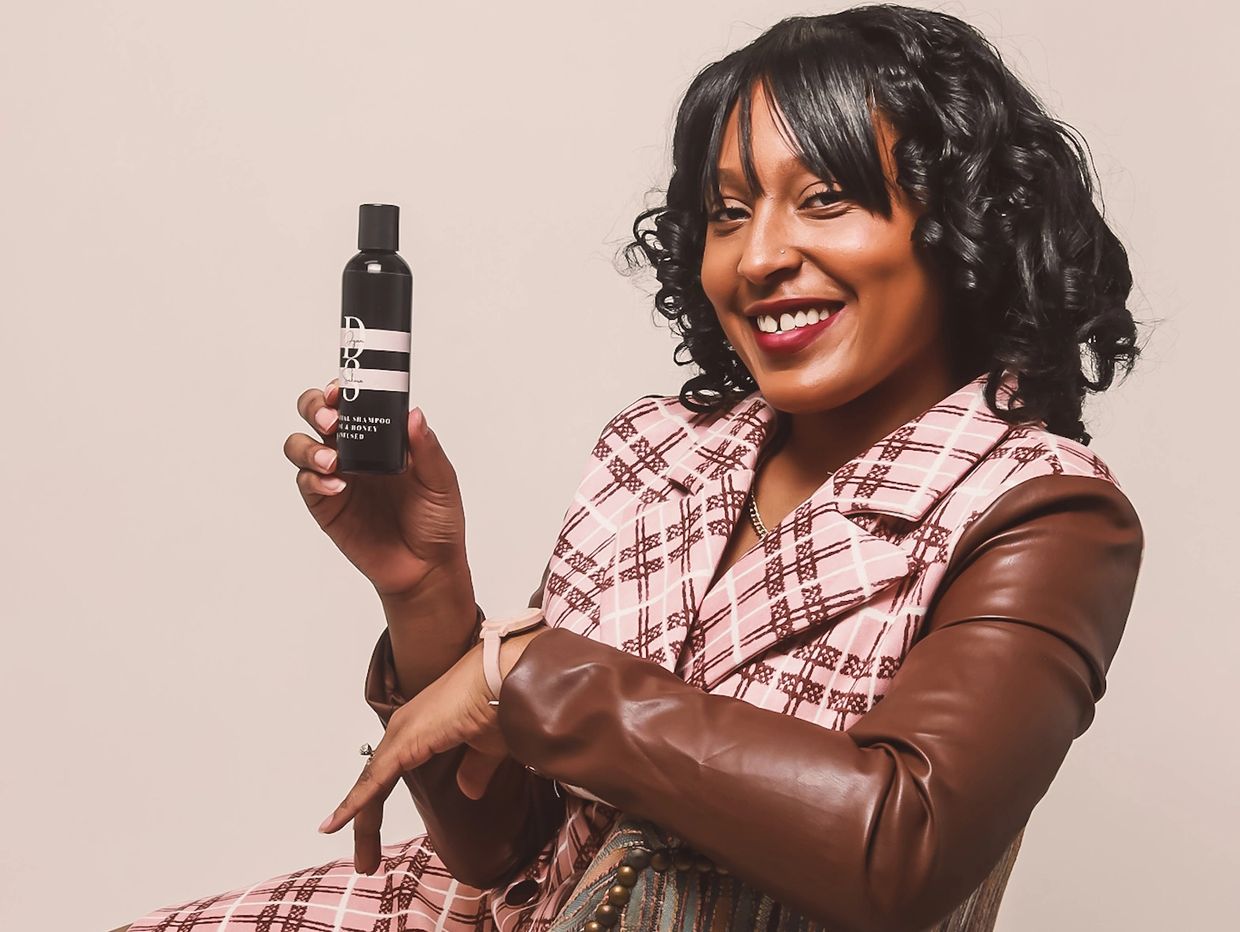 Smiling woman in stylish attire holding a black bottle product.