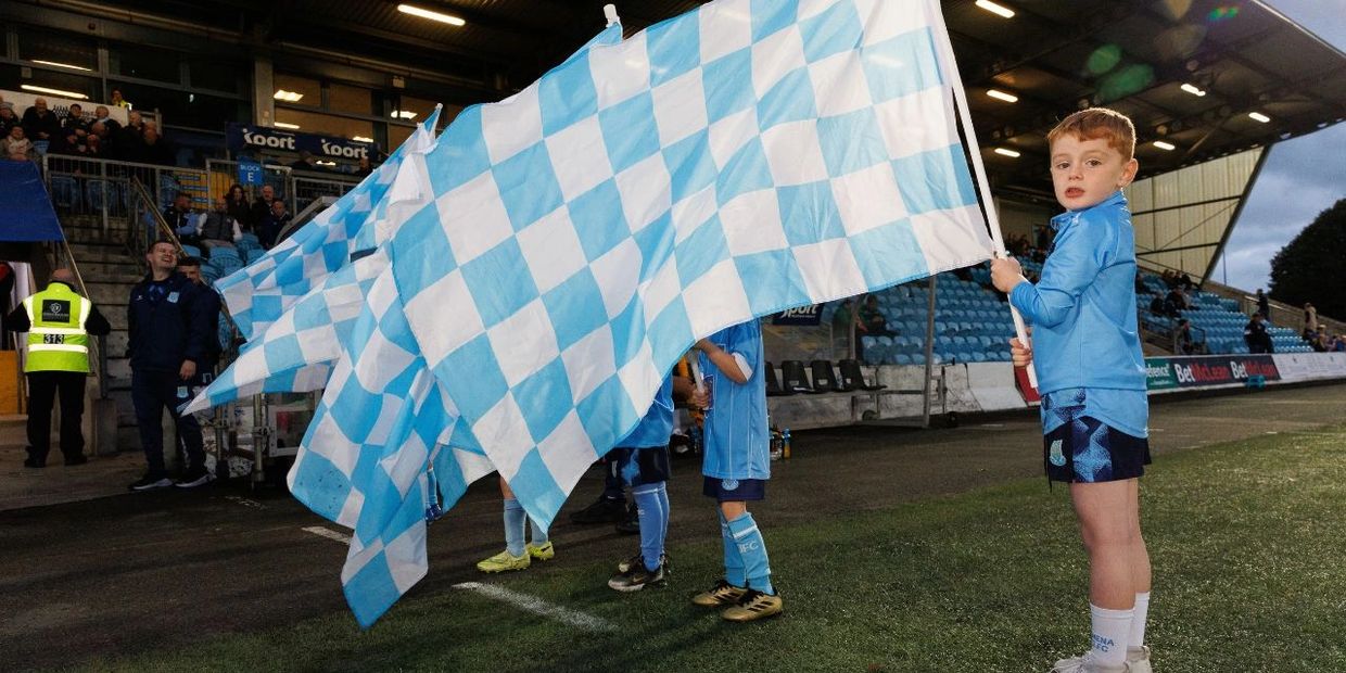 Young children holding blue and white checkered flags at a soccer stadium.