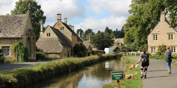 People walking dogs along a canal in a quaint village with stone houses.
