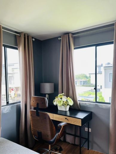 Cozy corner workspace with a wooden chair, black desk, and flower vase by large windows.