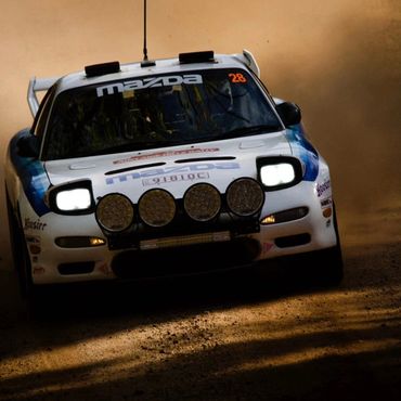 White Mazda rally car speeding on a dusty dirt road with headlights on.
