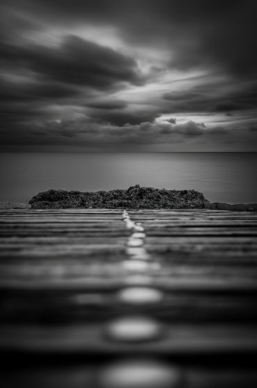 Black and white photo of a wooden pier leading to a rocky shore under a cloudy sky.