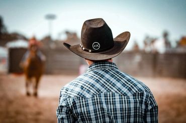 Back view of a cowboy wearing a hat and plaid shirt, with a horse rider in the background.