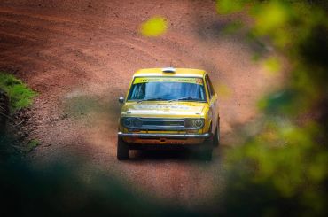 Yellow vintage rally car racing on a dirt track surrounded by greenery.