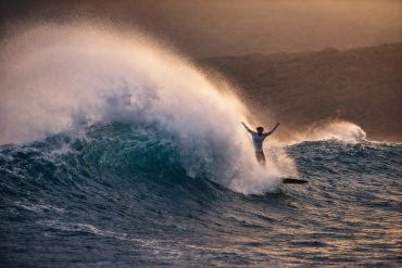 Surfer riding a large wave with arms raised during sunset.