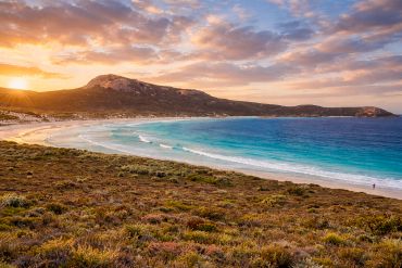 Sunset over a serene beach with turquoise waters and rocky hills.