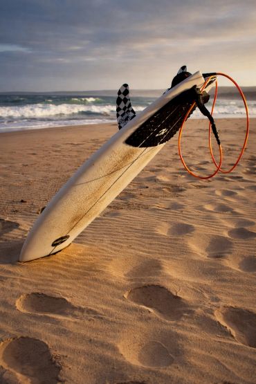 Surfboard stuck upright in sand at beach during sunset.