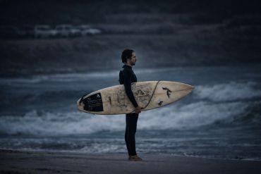 Surfer in wetsuit holding surfboard, standing on beach at dusk.
