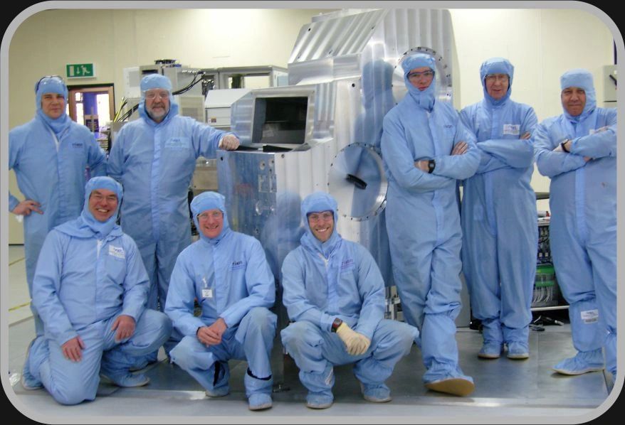 Group of people in cleanroom suits posing with industrial equipment.