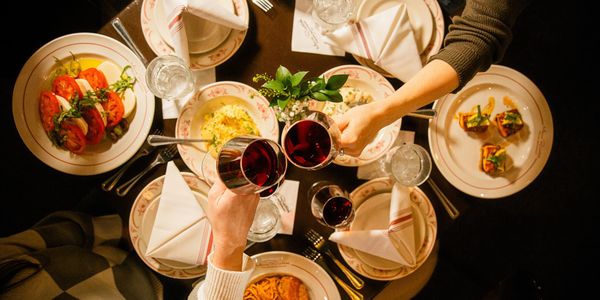 Two people clink glasses of red wine over a table with Italian dishes.