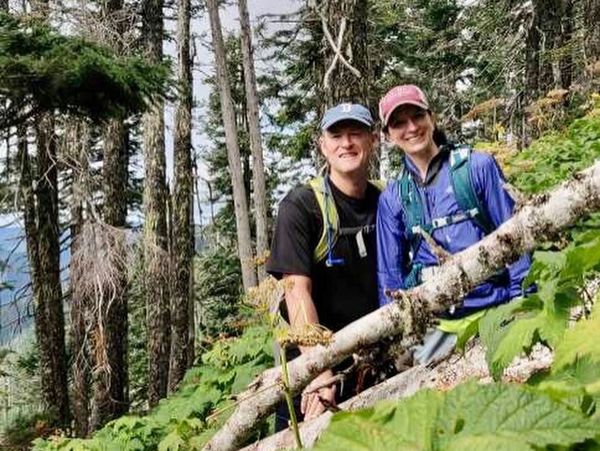 Two hikers smiling in a lush forest with backpacks and caps.