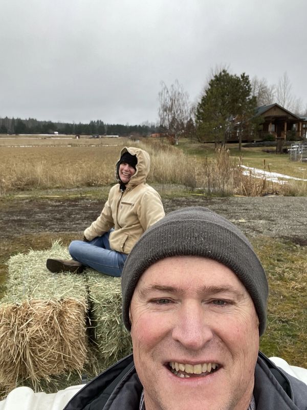 Two people outdoors on a cloudy day, one sitting on hay bales and the other smiling at the camera.