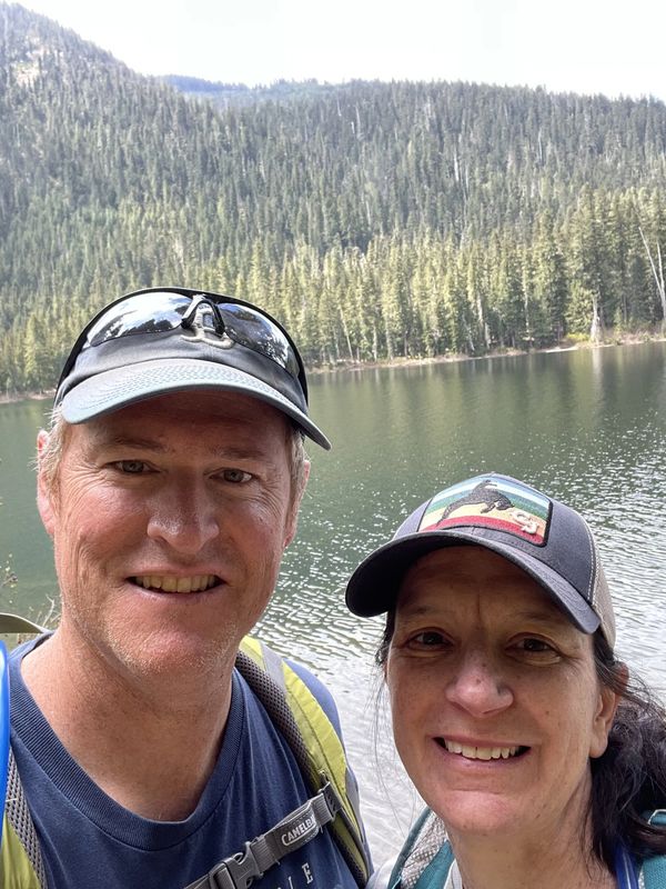 Couple smiling by a lake with forested hills in the background on a sunny day.