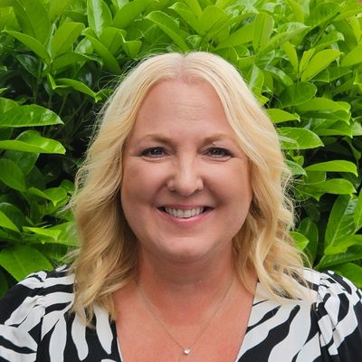 Smiling woman with blonde hair in front of green foliage.