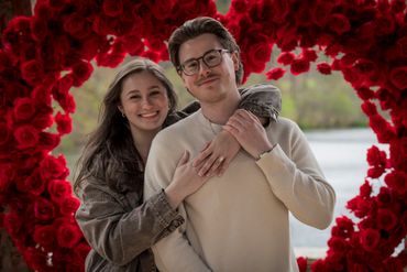 A couple embraces in front of a heart-shaped red flower backdrop.