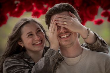 A woman playfully covers a man's eyes as they both smile under a red floral canopy.