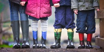 Four children standing in a row wearing rain boots, shown from the midline down