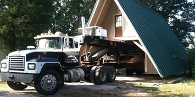 A truck moving an A-frame house in a wooded area.