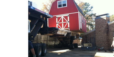 A red barn is being transported on a large truck near a house.