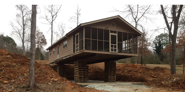 A house elevated on wooden supports in a clearing with bare trees.
