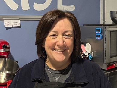 Smiling woman in a kitchen with utensils and appliances in the background.