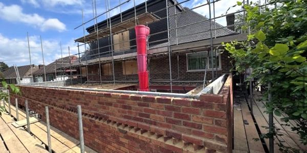 A brick building under construction with scaffolding and a red chute for debris.