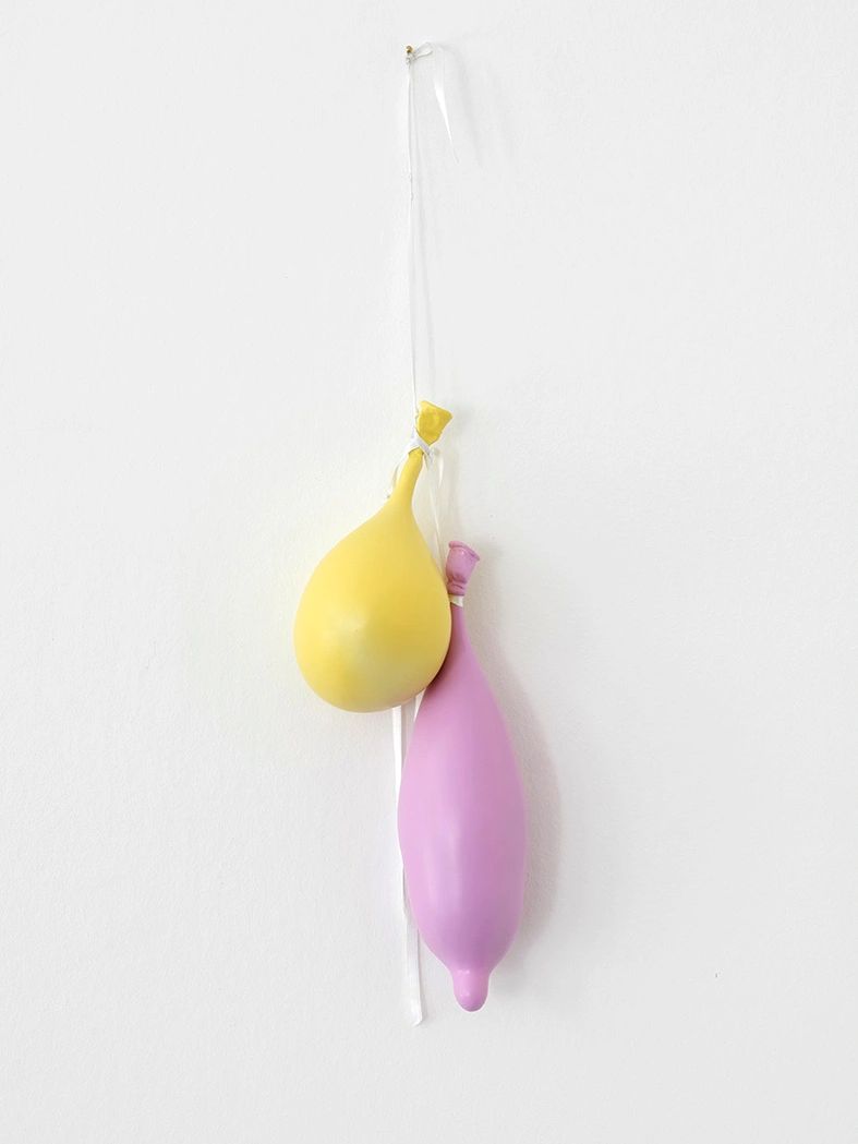 Two deflated balloons, one yellow and one purple, hanging against a white wall.