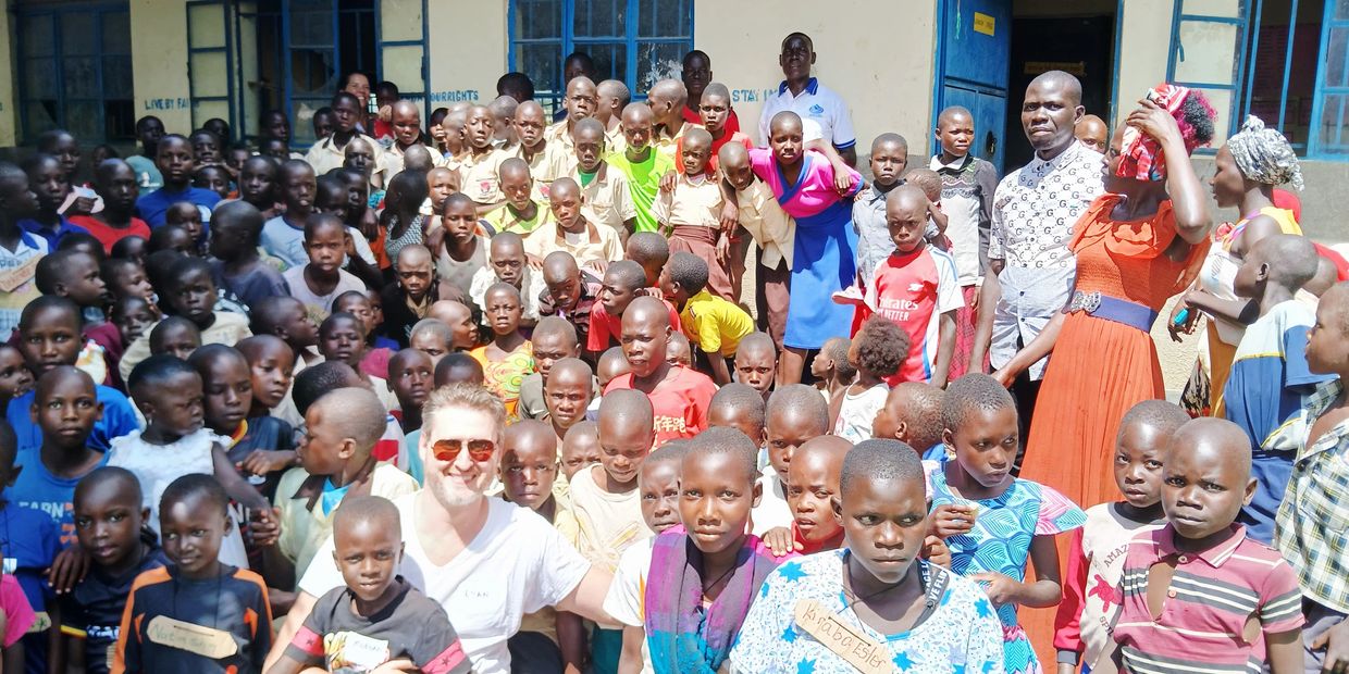 Large group of children and adults gathered outside a building, with one man wearing sunglasses in the front.