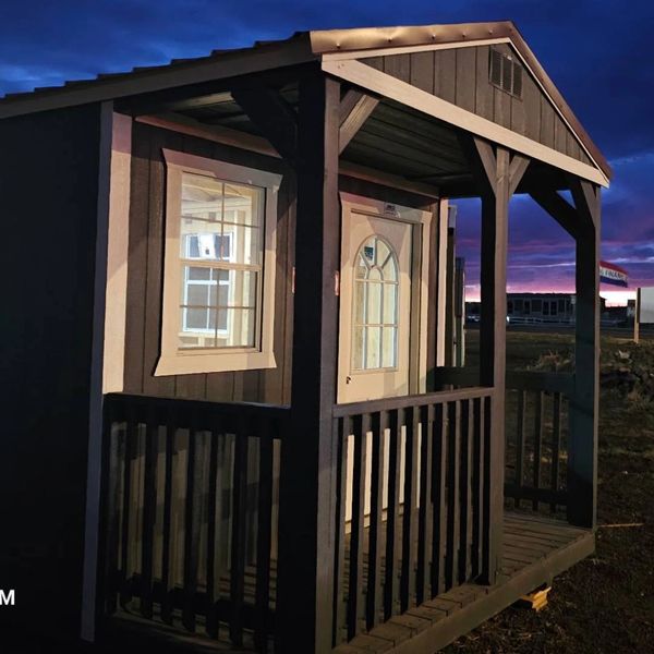 Portable shed with front porch and lit interior, shown at sunset – built by American Barn Co