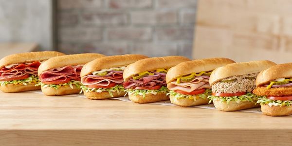 A lineup of seven assorted sub sandwiches on a wooden table.