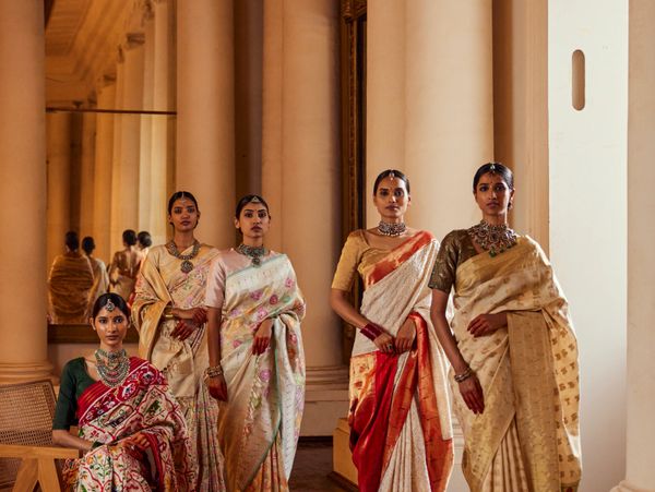 Five women dressed in elegant traditional sarees with jewelry in a grand hall.